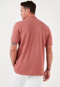 Lühikeste varrukatega polo särk tuharoosas, valmistatud pehmest kangast, millel on krae, kergelt tekstureeritud pind ja sirge alläär.