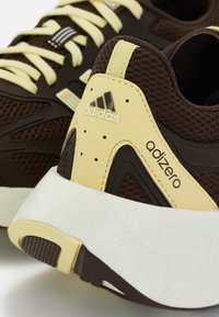 Rudos tinkliniai kedai su geltonais akcentais. Yra Adidas logotipas, tekstūruotas vir�šus ir balta pada su išskirtiniu dizainu ir raštu.