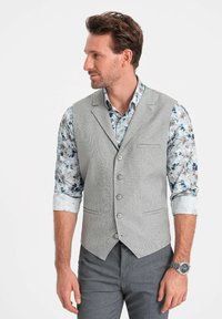 Gray textured vest with five buttons, paired with a floral patterned shirt. Model wearing gray trousers and a watch. Side profile view.