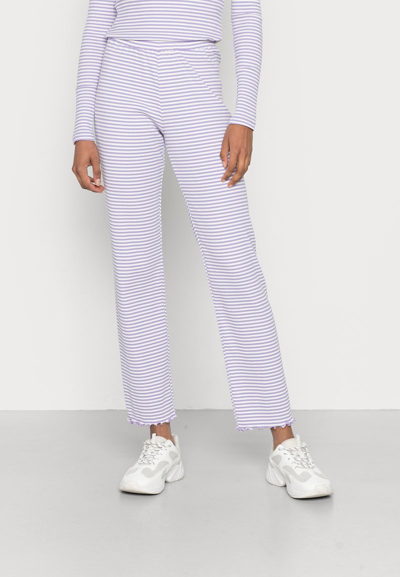 Calças listradas em roxo e branco com um corte solto, apresentando uma bainha desfiada e combinadas com ténis atléticos brancos.