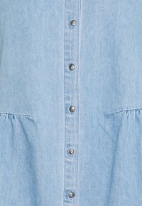 Tela de mezclilla azul claro con una fila vertical de botones de metal plateado y costuras sutiles fruncidas a ambos lados debajo de los botones.