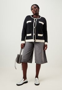 Svart og kremfarget cardigan med knapper og to frontlommer, sammen med grå denimshorts og sølvfargede slip-on sko. Mønstrete veske som tilbehør.