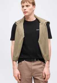 Νεαρός άνδρας που φοράει μαύρο t-shirt BOSS με ένα ελαιί πράσινο πουλόβερ περασμένο στους ώμους και καφέ παντελόνια, στέκεται μπροστά σε απλό φόντο.