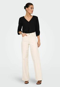 Schwarze V-Ausschnitt-Bluse mit ¾-Ärmeln, kombiniert mit hochtaillierten, weit geschnittenen cremefarbenen Hosen. Das Modell trägt schwarze Riemchensandalen und silberne Accessoires.