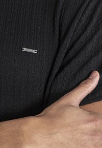 Czarny krótki rękaw top o fakturowanej powierzchni, z wzorem w warkocze i srebrną metką marki Kavanagh po lewej stronie bluzki w okolicy dekoltu.