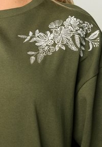 Felpa verde oliva con ricami floreali bianchi sulla spalla, texture morbida e vestibilità rilassata con scollatura rotonda.