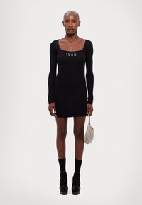 Black long-sleeve dress with a square neckline, featuring the word "ICON" in white. Accessories include a metallic handbag and platform shoes.