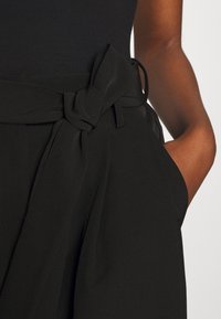 Pantalon noir taille haute avec une large ceinture en tissu nouée en nœud, porté par une personne avec la main droite dans la poche.