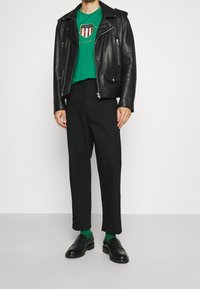 Czarna skórzana kurtka motocyklowa na zielonym t-shircie z grafiką, w połączeniu z czarnymi spodniami i czarnymi loafersami. Widoczne zielone skarpetki.