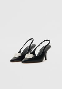 Musta slingback-korkokengät, jossa on terävä kärki ja sydämenmuotoinen strassikoriste kengän päällä, sileä pinta ja hoikka stiletto-korko.
