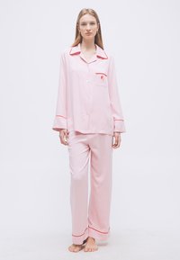 Γυναίκα που στέκεται ξυπόλητη φορώντας ανοιχτό ροζ πιτζάμα με μακριά μανίκια, με κόκκινα περιγράμματα και μικρό κεντητό λογότυπο στην τσέπη του στήθους.