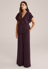 Top in lino viola con maniche corte e cintura annodata, abbinato a pantaloni a gamba larga coordinati, caratterizzati da un design fluido e una texture morbida.