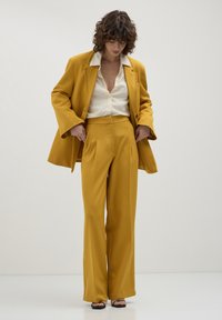 Abito su misura giallo senape con pantaloni a gamba larga e blazer coordinato. L'outfit presenta una camicia bianca sotto.