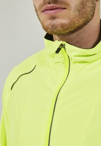 Veste de sport jaune fluo avec un col haut, dotée d'une fermeture éclair et de accents noirs contrastants le long des coutures pour plus de détails.