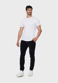 T-shirt blanc à manches courtes, pantalon noir ajusté et baskets blanches. Le mannequin se tient de manière décontractée avec une main dans sa poche, design et couleurs minimalistes.