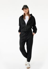 Conjunto de sudadera con capucha negra y joggers con un acabado texturizado, que cuenta con un cierre frontal de cremallera y cintura con cordón, combinado con una gorra negra y zapatillas blancas.