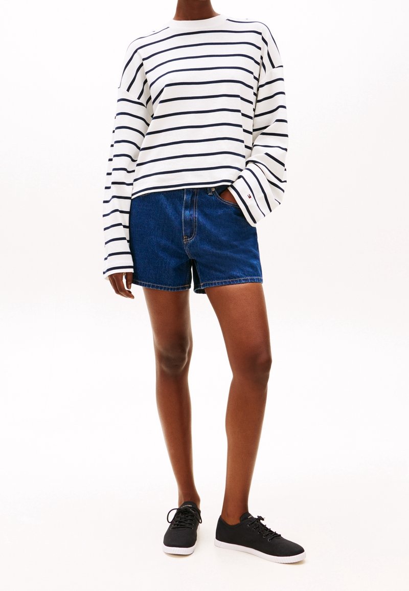 Personne portant une chemise blanche à manches longues avec des rayures horizontales noires, un short en denim bleu foncé et des baskets noires avec des semelles blanches.