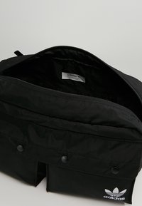 Svart Adidas ryggsäck med öppen huvuddragkedja, två framfickor med tryckknapp och vit Adidas-logotyp på framfickan.