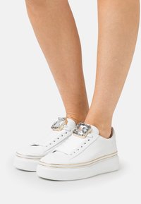 Sneakers bianchi in pelle con suola piatta, caratterizzati da grandi accenti di cristallo decorativi sui lati esterni e rifiniture dorate attorno alla suola.