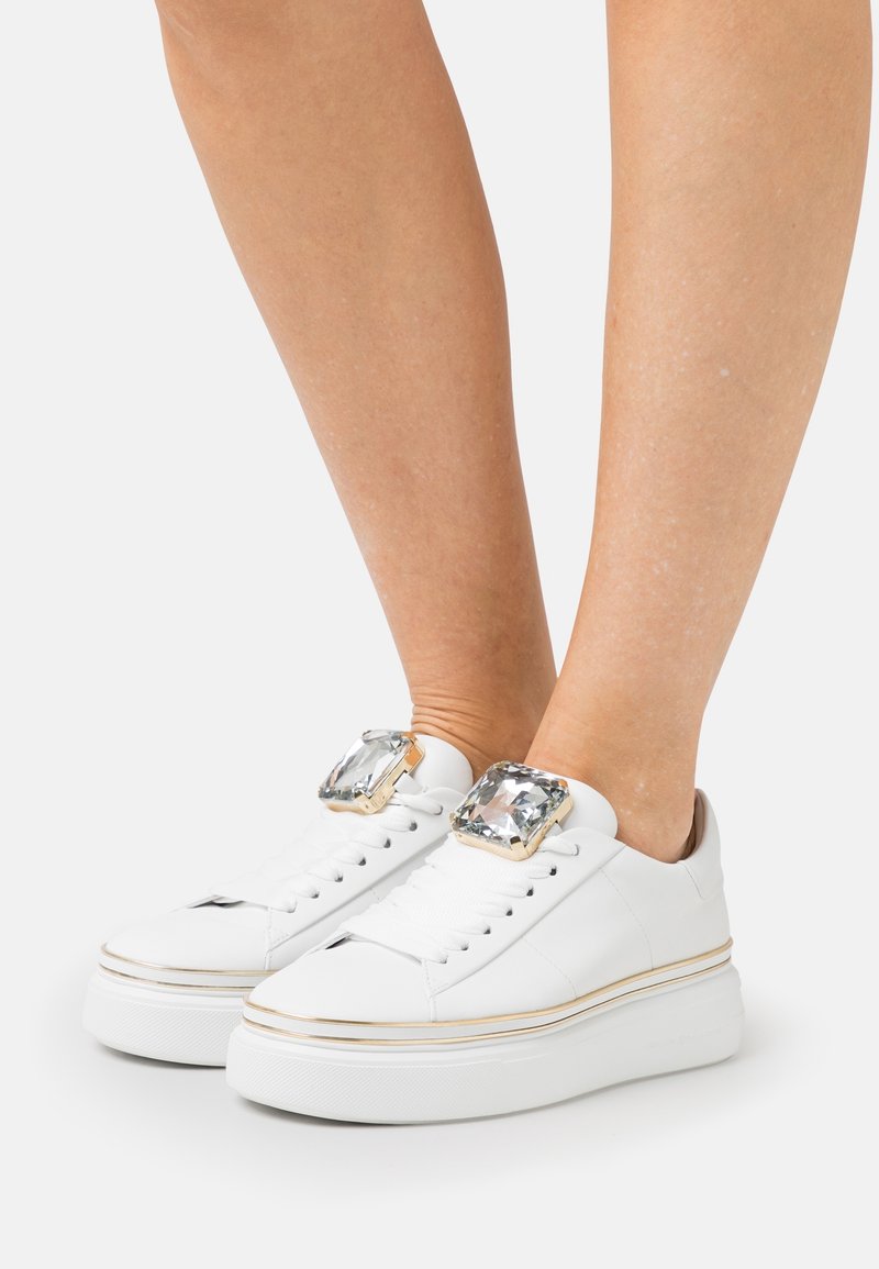 Sneakers bianchi in pelle con suola piatta, caratterizzati da grandi accenti di cristallo decorativi sui lati esterni e rifiniture dorate attorno alla suola.