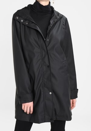 Long imperméable noir avec capuche porté par-dessus un col roulé noir et un pantalon noir, boutons et fermeture éclair visibles à l'avant, mains détendues le long du corps.