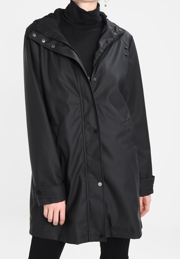 Long imperméable noir avec capuche porté par-dessus un col roulé noir et un pantalon noir, boutons et fermeture éclair visibles à l'avant, mains détendues le long du corps.