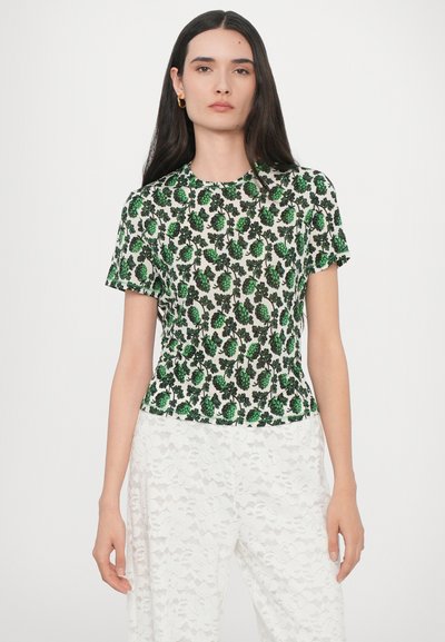 Top corto a maniche corte con stampa floreale verde e scollatura rotonda, realizzato in un tessuto leggero. Indossato con pantaloni bianchi in pizzo.