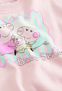 Sudadera rosa con cerditos de dibujos animados. El diseño incluye tres personajes sobre un fondo crema y acentos a rayas turquesas. Texto "Hermana Pequeña".