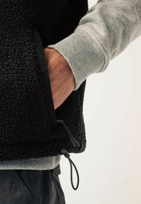 Czarna kamizelka polarowa o teksturowanej powierzchni, z szarym swetrem jako warstwą dolną i regulowanym sznurkiem ściągającym u dołu. Widać kieszeń.