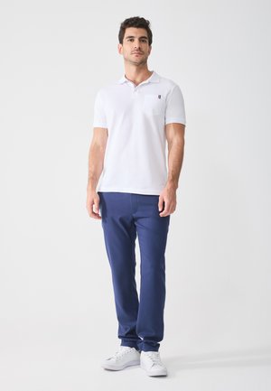 Chemise polo blanche à manches courtes et avec une poche, associée à un pantalon slim fit bleu et des baskets blanches. Conception simple, sans motifs ni accents.