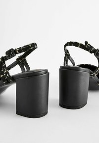 Sandales noires à talons blocs avec des brides en tissu texturé et des boucles ajustables, vues de dos sur un fond blanc.