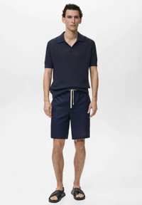 Polo shirt navy a maglia con maniche corte, abbinato a pantaloni corti navy con vita regolabile e indossato con sandali neri a ciabatta.