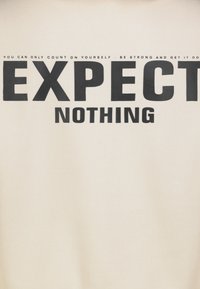 Cremefarbenes Gewebe mit fettem schwarzem Text. Der zentrale Text zeigt "EXPECT" über "NOTHING", mit kleinerem Text darüber. Weiche Textur.