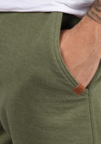 Pantalons de jogging vert olive en matériau doux, avec des poches latérales et une étiquette en cuir près de la couture de la taille.