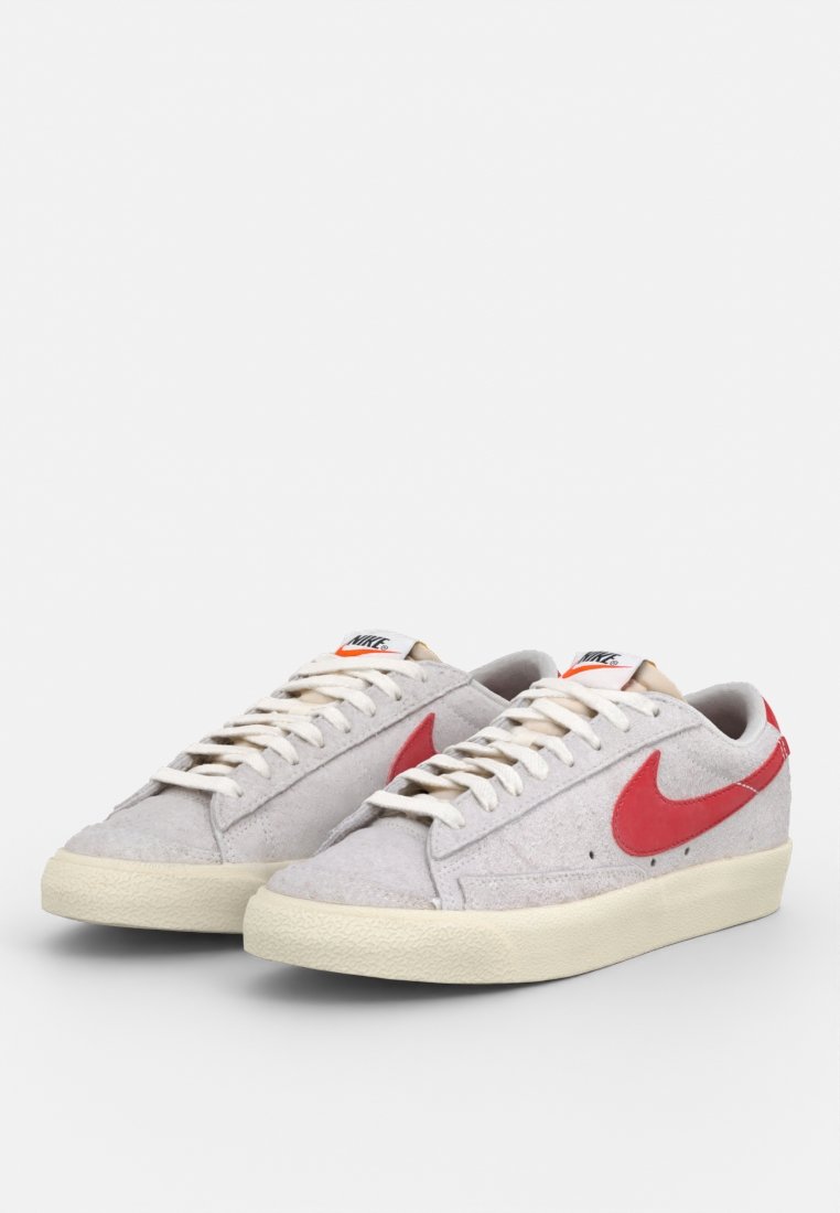 Sneaker in scamosciato grigio con logo Swoosh rosso, suola in gomma crema e lacci beige. Presenta un'etichetta Nike sulla linguetta.