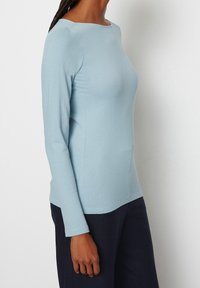 Ljusblå långärmad topp med textilstruktur, vid halsringning och formad silhuett, i kombination med mörka byxor.
