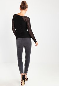 Schwarzes Langarmshirt mit Spitzenverzierung und strukturiertem Vorderteil, kombiniert mit grauen engen Jeans mit Reißverschlüssen an den Knöcheln und hochhackigen Sandalen.