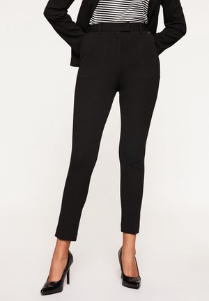 Vrouw die zwarte, nauwsluitende enkellange broek draagt, een zwarte blazer, een gestreept overhemd en zwarte hoge hakken, staand met haar handen op haar heupen.