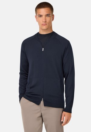 Navy zip-up cardigan made of smooth fabric, featuring a ribbed collar, long sleeves, and two front pockets; fitted design with a clean finish.