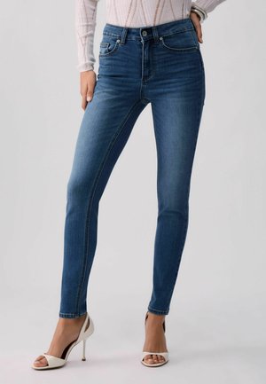 Donna con jeans skinny blu a vita media e sandali bianchi aperti con tacco alto, mano appoggiata sull'anca, su sfondo neutro.