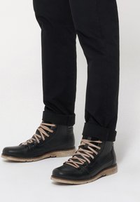 Czarne skórzane buty z jasnobrązowymi sznurówkami, teksturowana powierzchnia i kontrastująca jasnobrązowa podeszwa. Spodnie podwinięte na kostce.