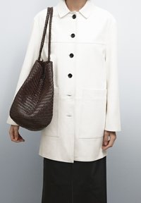 Cream-colored, button-up coat with black buttons, two front pockets, paired with a woven, dark brown tote bag. Smooth fabric texture.