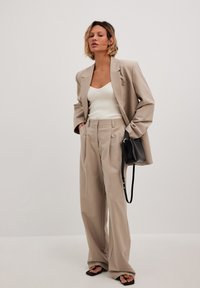 Beige skräddarsydd kavaj, ribbad vit linne, och löst sittande beiga byxor. Svart handväska med rem och sandaler med öppen tå.