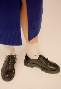 Scarpe in pelle nera con punte tonde e suole piatte. Presentano dettagli con allacciatura e sono abbinate a calzini chiari.