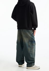 Svart huvtröja i oversized modell, tillsammans med oversized blå denim cargobyxor som har flera fickor och ljusa tvättad detaljer. Vita sneakers.