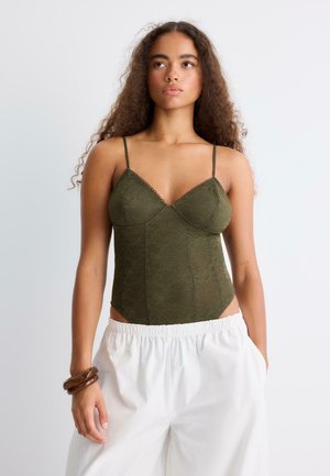 Femme aux longs cheveux bouclés porte un caraco en dentelle vert olive et un pantalon blanc taille élastique, debout sur un fond clair, les mains dans les poches.