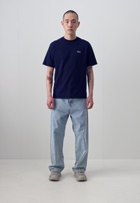Tricou din bumbac bleumarin cu un logo mic pe piept, asociat cu blugi cu spălare deschisă și pantofi sport gri. Fundal simplu.