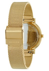 Montre avec bracelet en maille dorée et boîtier rond minimaliste. Présente un fond en verre lisse et réfléchissant ainsi qu'un fermoir sécurisé avec logo gravé.