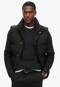Chaleco de plumas negro con cuello alto y botones a presión, usado sobre un suéter de punto oscuro. Tejido texturizado, parte delantera cerrada, detalles de costura prominentes.