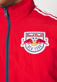 Rød zip-jakke i lyse røde stof med hvide striber. Har et broderet Red Bull New York-logo på venstre bryst.